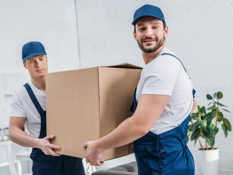 two-handsome-movers-transporting-cardboard-box-in-apartment-e1620131893815.jpg